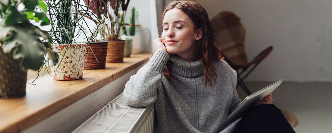 Jonge vrouw zit genietend bij de verwarming. In de vensterbank staan planten | verwarm je huis veilig | a.s.r.