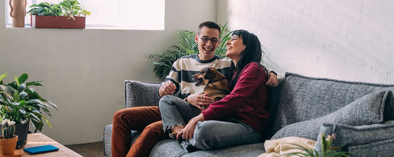 Jonge man en vrouw zitten lachend op de bank met hun hondje in hun handen