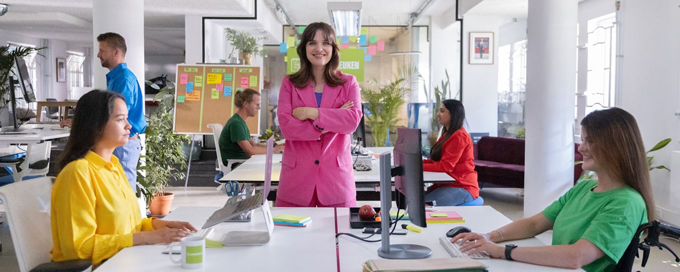 Vrouw staat in roze pak met armen over elkaar aan bureau. Vier andere mensen in gekleurde kleding zitten achter hun bureau achter een computer